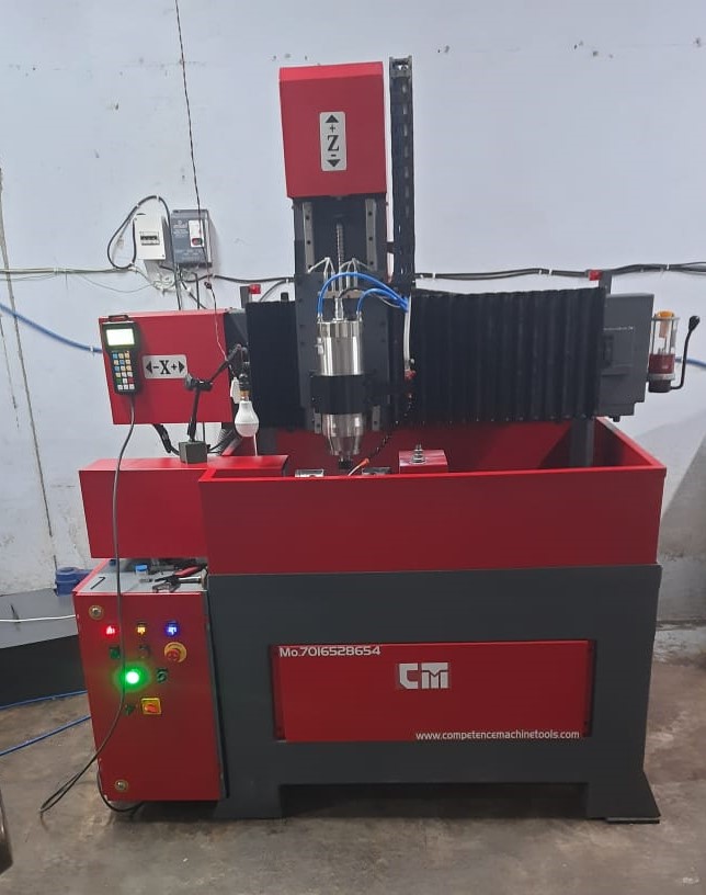 A red and gray industrial CNC machine on a concrete floor. It has control buttons and display screens, with metal tools and wires visible.