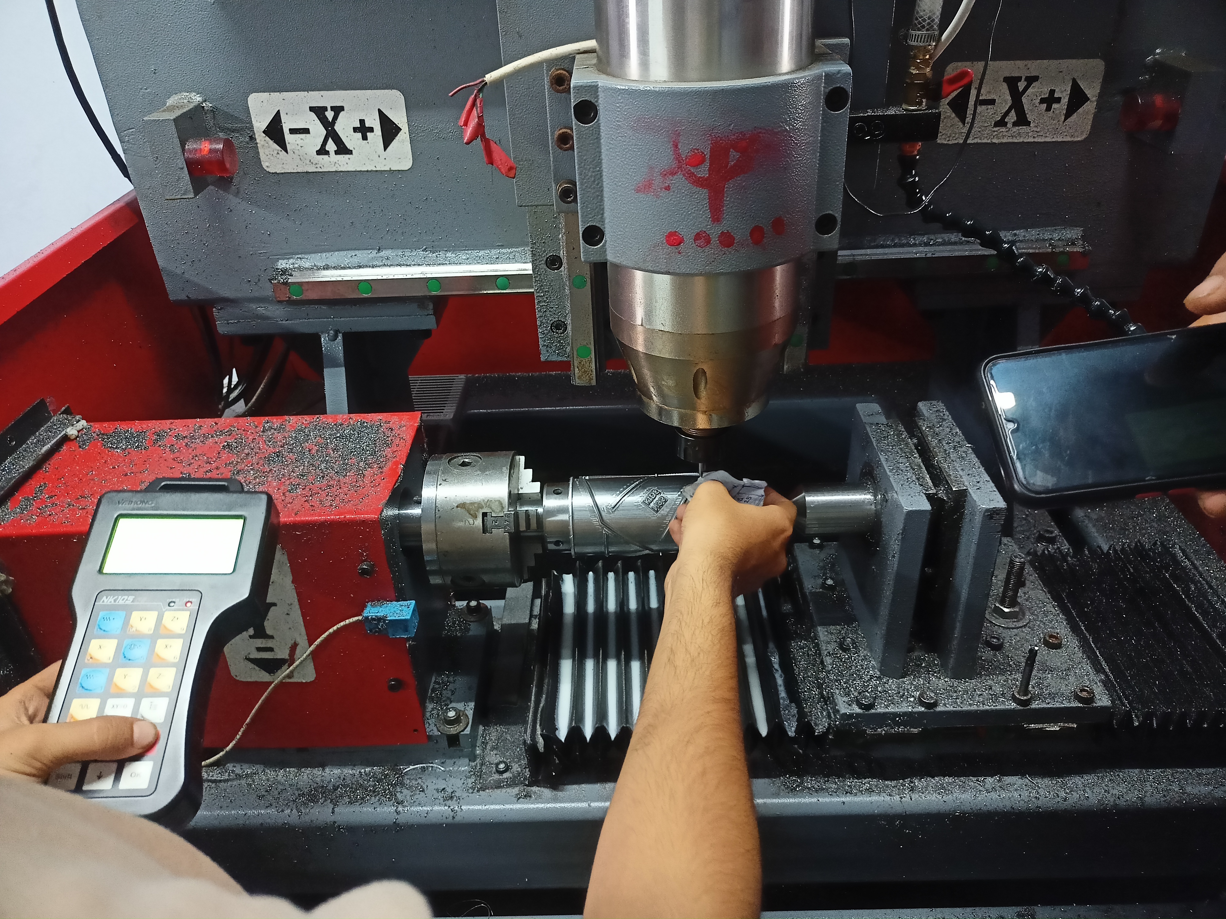 A close-up of a CNC machine in action. A person's hand adjusts a cylindrical metal part. One hand holds a control device, another holds a smartphone, capturing the process. The machine is detailed, with metal shavings around it, conveying a technical and industrious atmosphere.
