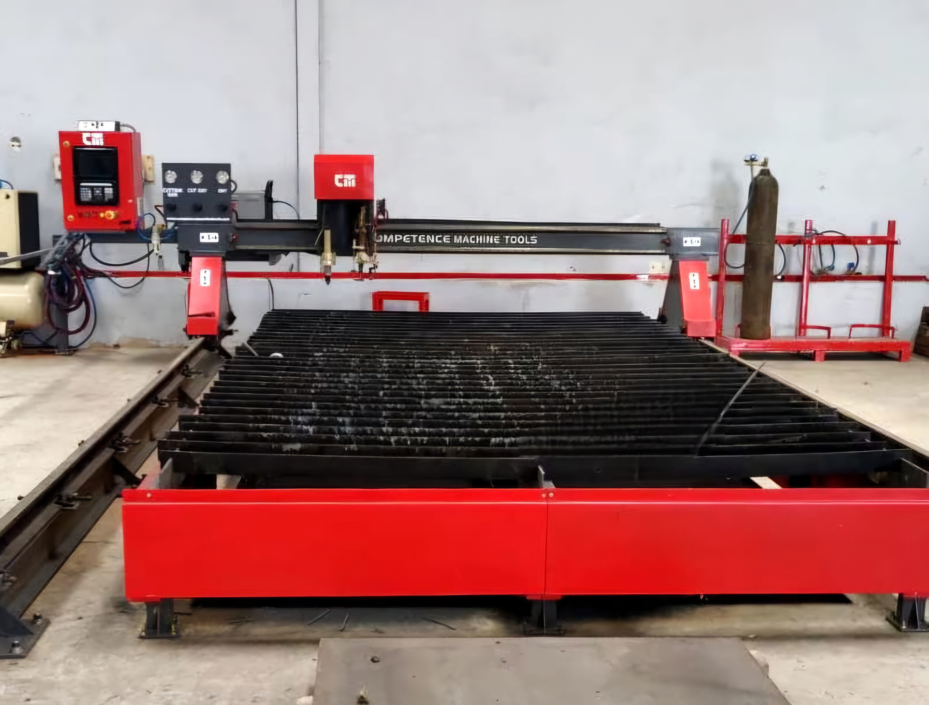 Large industrial CNC plasma cutting machine in a workshop, featuring red and black components, control panel, and gas cylinder, conveying precision engineering.