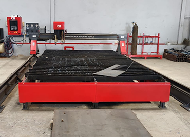 Industrial machine in a workshop with a large red base and black grate surface. Control panels and metal cylinders are visible in the background.