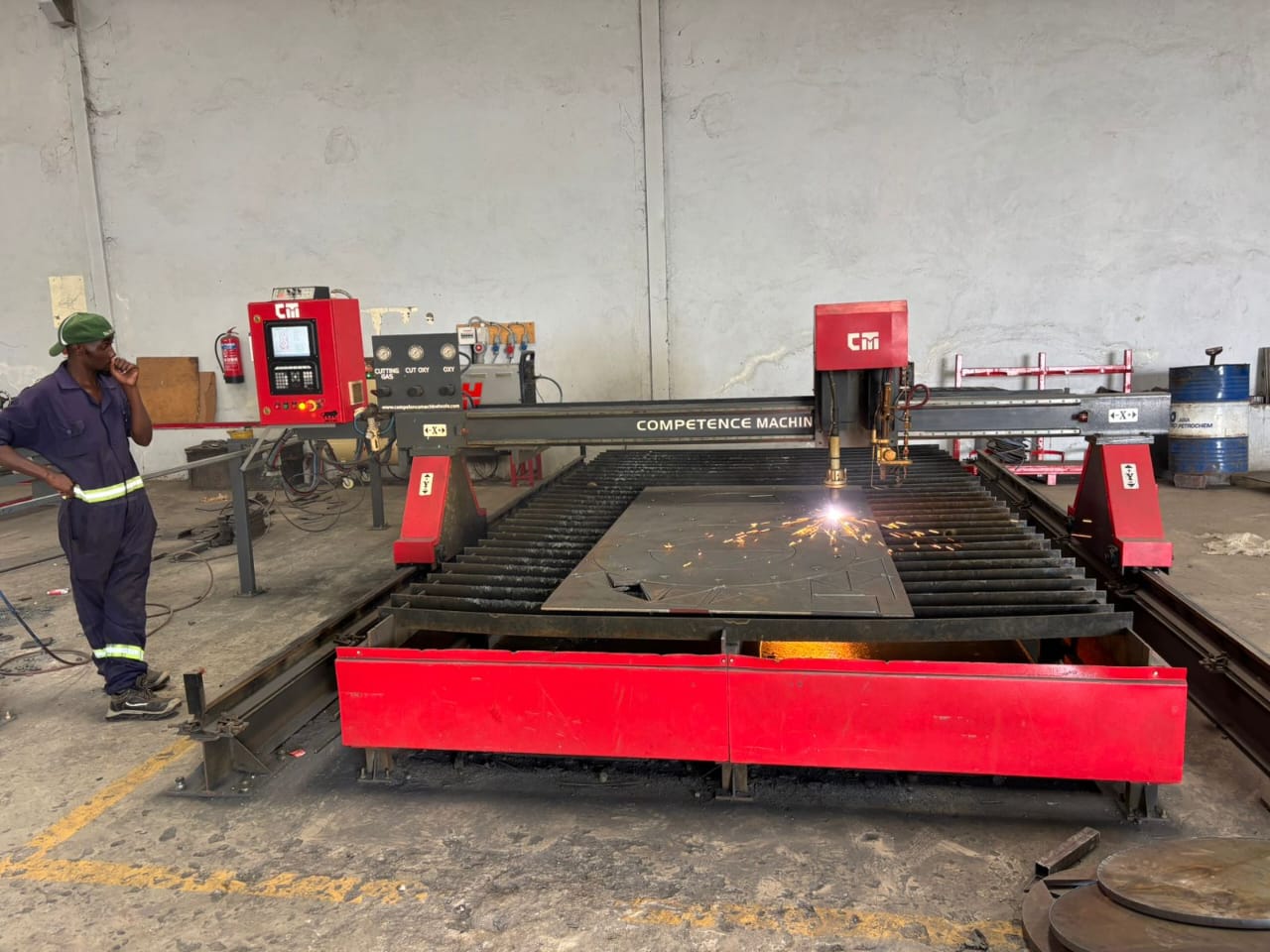 A man in a workshop observes a large red CNC machine cutting metal, with sparks flying. The atmosphere is industrious and focused.