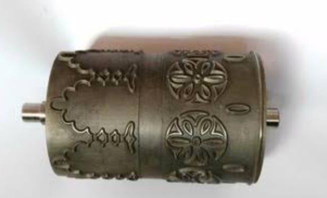 A cylindrical metal embossing roller featuring intricate floral and geometric patterns. Its surface is textured, indicating a decorative purpose.