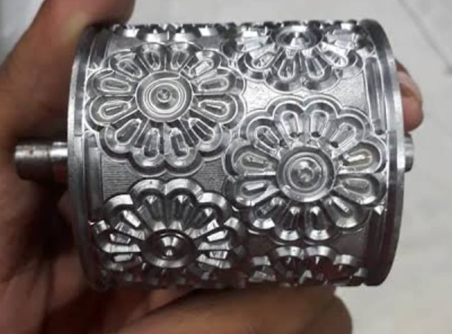 Close-up of a metallic cylinder with intricate floral engravings, held by a hand. The shiny surface reflects light, highlighting the detailed patterns.